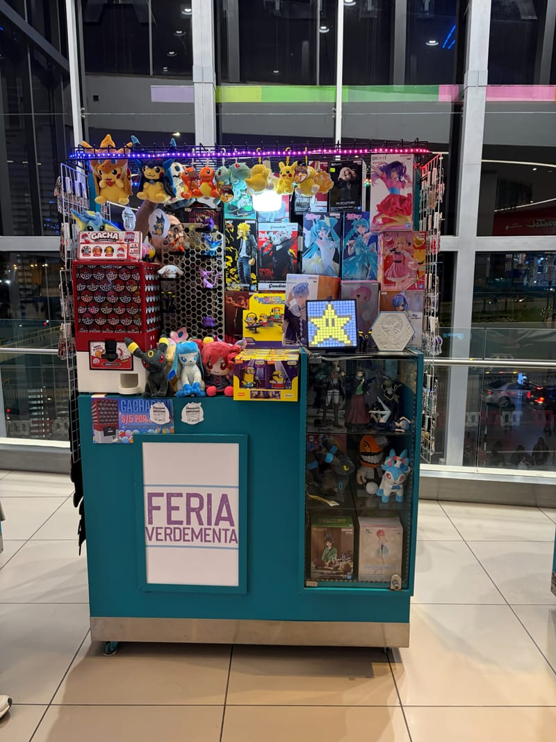 Nuestro stand en la Feria Verdementa: mostrador turquesa con el cartel de Feria Verdementa, peluches de Pokémon colgados arriba y figuras de anime en la vitrina.